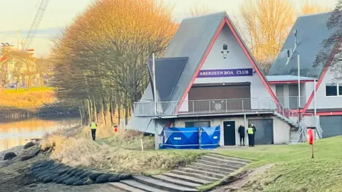 A blue police tent pitched up in front of a triangular shaped building which has a sign on it saying Aberdeen boat club. Police officers can be seen surrounding it.