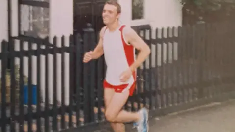 GRAHAM BREW An old photo of Graham Brew, who is smiling while running. He is wearing red and white running gear and is running past a house with a large black fence.