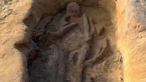 Oxford Cotswold Archaeology Shadow skeletons of an adult and child emerging from sandy soil in a pit. The head of the adult is at the top and its arms, torso and legs can be seen. The bod of the child is tucked in at its right side, with its left arm folded over its body.