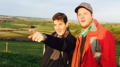 Dr Tom Pelly and his brother. Dr Pelly is pointing at something in the distance. They are in a remote location, with green hills and fields behind them.