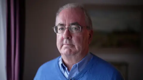 An older man with glasses, wearing a blue v neck jumper and a blue collar shirt stares at the camera. His face is lit by light from a window to his right.