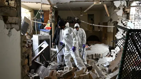 Getty Images Forensic officials use metal detectors to inspect amid the debris of a damaged house after a gas cylinder explosion in a Christian colony in Islamabad on January 11, 2026