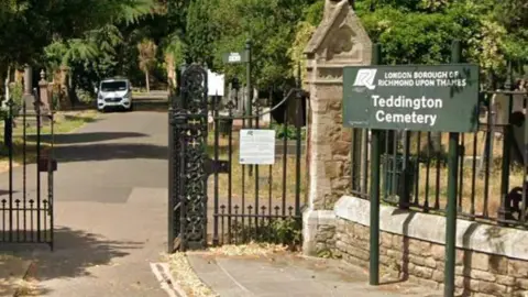 The stone front entrance and iron gates of a cemetery - to the right is an elevated sign that says London Borough of Richmond upon Thames, Teddington Cemetery. 