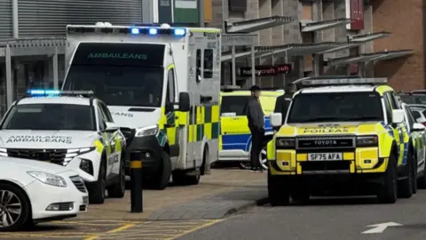BBC Police Scotland and Scottish Ambulance Service parked next to a row of shops at Inverness' Inshes Retail Park.