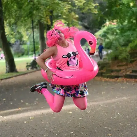 Jo Yarnall A woman jumps in the air in pink clothing, with a pink inflatable flamingo ring around her waist