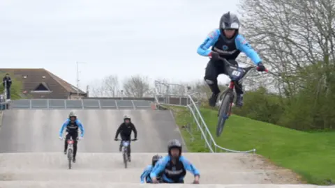 A group of BMX riders hurtling down a track. There are five of them each on their bike in the picture. At the front one of the riders is going over a bump on the track and has been propelled into the air.