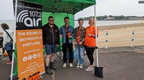 Two men and two women stand smiling underneath a gazebo next to a sign for the Air radio station. They're on a promenade with a beach behind them.