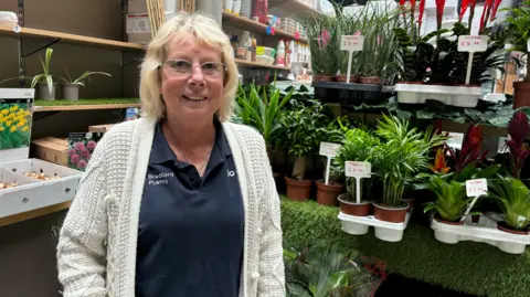 A woman with blonde hair and glasses smiles at the camera. She is wearing a cream, knitted cardigan and a navy blue polo shirt with Bradford Plants written on it. To her right is a selection of green pot plants.