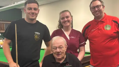 Simon Thake An elderly man in a wheelchair sits flanked by two men and a woman who stand behind him. In the background is a snooker table