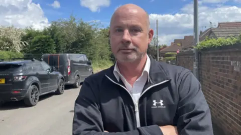 A mid shot of a man with no hair, folding his arms and looking into the camera. He is wearing a black jacket. There are several cars behind him.