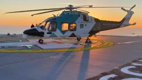A blue and white helicopter on a helipad, with the letters RSCH painted on the surface. An orange sunset and a view over the city of Brighton can be seen in the background.