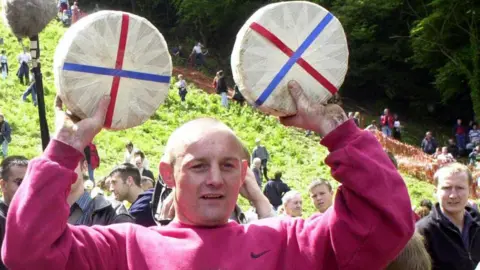 A man with a light coloured buzzcut holds two wheels of cheeses at the foot of Cooper's Hill near Gloucester following the annual cheese rolling races. He is wearing a fuchsia coloured Nike sweatshirt. The steep hill can be seen behind him, as can a crowd of people.