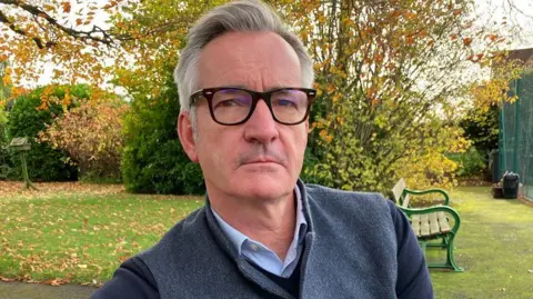 Robert Marsh sits on a bench in a park setting with a tree and grass behind him and he is looking into the camera 