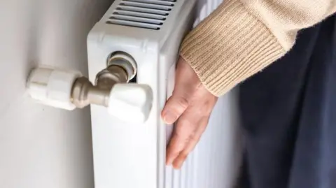 A woman touches a radiator in a house to check how warm it is. She is wearing a beige jumper. The close-up picture shows her hand and sleeve.