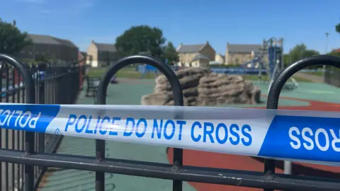 A close-up image of blue and white police tape with the words do not cross surrounding a playground with houses in the distance