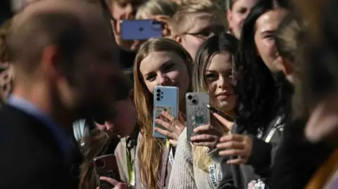 PA Media A crowd of young people pointing their cameras to a blurred image of a man in a dark jacket and blue shirt.