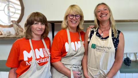 Three women stand in a row - Elaine and Linda, to the left and the centre, both wear orange MND association t-shirts and Sarah (to the right) is in a polkadot t-shirt. All wear beige aprons.