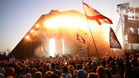 Reuters A crowd is watching Kendrick Lamar performing at Glastonbury 2024. The stage lights are golden and shining onto the crowd. People are holding up some flags.