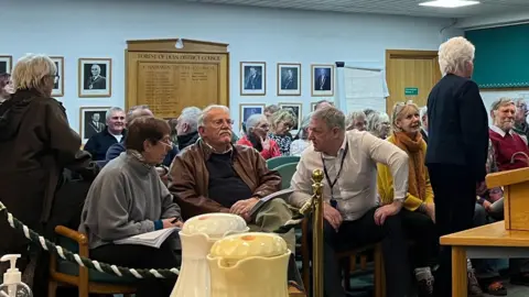 LDRS Residents fill out a public gallery inside a council chamber, the walls of which are decorated with portraits of previous chairmen and a large wooden plaque, commemorating their names. The residents, all older people, are sat on chairs, talking to each other, with a few people seen making their 