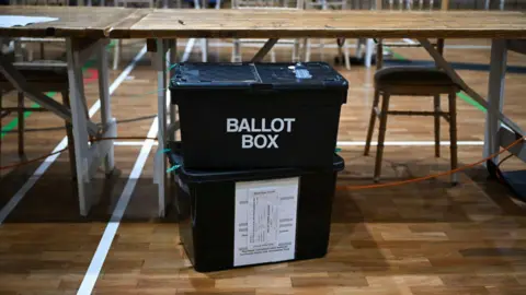 A stock image showing two black ballot boxes stacked on top of each other in a large gym. Behind it are rows of wooden chairs and tables for counters to use.