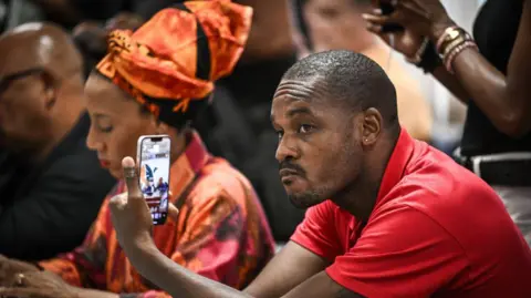 PHILIPPE LOPEZ/AFP Rodrigue Petito, leader du Rassemblement pour la protection des populations et des ressources afro-caribéennes (RPPRAC), vêtu d'un t-shirt rouge, regarde son téléphone portable, lors d'une réunion. 