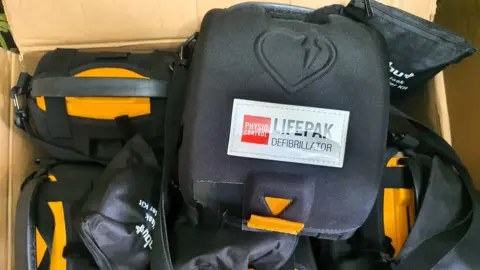 A cardboard box of multiple defibrillators in black bags with yellow features and a label reading 'physio control, lifepak defibrillator'.