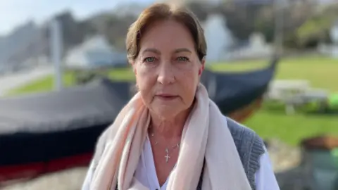 Julie Pinson has blue eyes and brown hair tied up. She is wearing a neutral pashmina over a white. shirt and grey cardigan. She is standing in front of a covered dinghy on a sunny day.