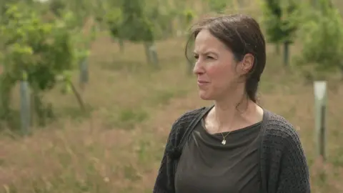A woman in a dark top and cardigan with long dark hair and wearing a gold necklace looks to the left. Behind her is an area of long grass with young trees planted on it. They still have their protective plastic wrappers around the trunks.