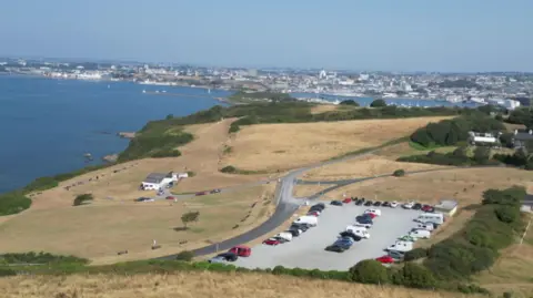 A drone shot of Jennycliff car park taken on a bright clear sunny day in Plymouth. The car park is visible as well as scorched yellow grass and the city and Plymouth Sound in the background.