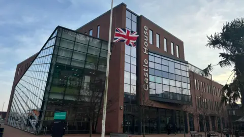 A large brick and glass building bearing the words Castle House. In the foreground, the red, white and blue union flag is flying at half mast.