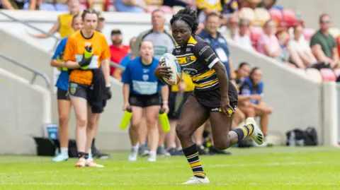 Craig Hawkhead Photography Peace Lekuru in action on the rugby pitch, running with the rugby ball. She wears a York Valkyrie rugby shirt which is black with a yellow stripe across the chest, black shorts, and black and yellow striped socks. Spectators can be seen behind her in the stands, and teammates watch on.