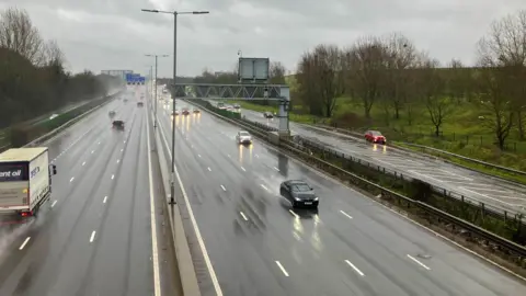 s Pace Both lanes of the motorway, with cars creating splashes of water as they drive across the wet surface. The picture is likely taken from a bridge.