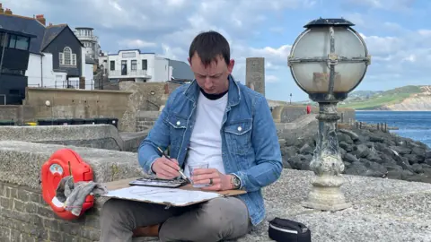 A man in a blue denim jacket sits on a wall with the sea and Golden Cap in the background. He's got a wooden drawing board on his lap and is painting a water colour picture of a dinosaur/