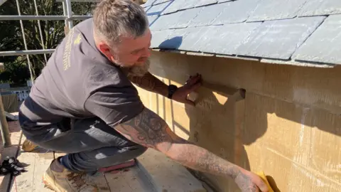 a tattooed man wearing a t-shirt sponges down sandstone while standing on scaffolding at the church. You can faintly see an engraving on one fo the stones saying High Toynton 2025