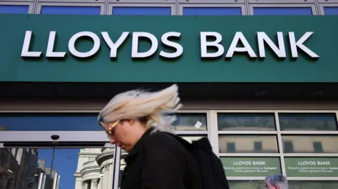 The Lloyds Bank logo on the outside of a high street bank. In the foreground a woman with blonde hair walks past