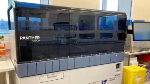 A polymerase chain reaction machine, a square box which has a grey plastic window, and the words "panther fusion" on it.