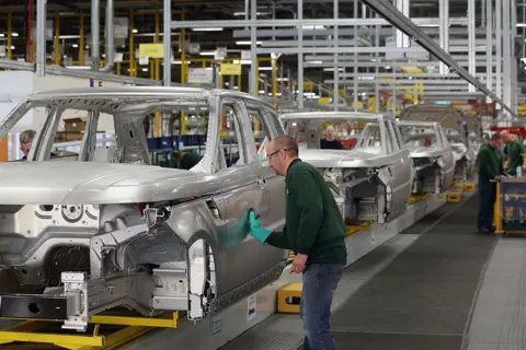 A factory line of shells of cars- a man wearing a green jumper, safety goggles and a green glove is standing sideways working on one of the cars. Colleagues can be seen in the background also wearing green jumpers. 