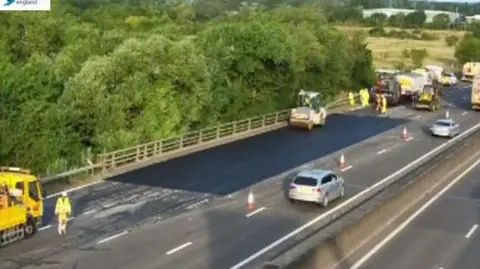 A blurry traffic camera image showing the M25 near junction 25. Traffic is down to one lane and going past a patch which has fresh black tarmac on it. There are emergency vehicles in yellow and workers also in hi-vis yellow clothing. 