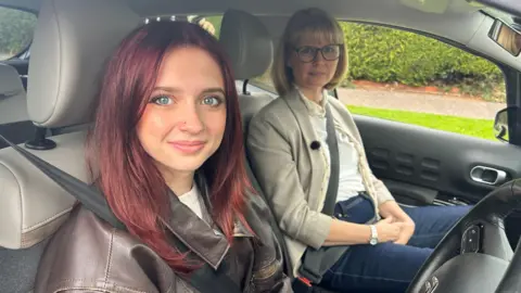 BBC A girl with red hair wearing a brown leather jacket sits next to her mum who has short bobbed blonde hair in a car. They are both looking at the camera.