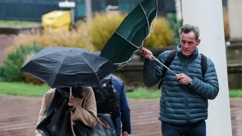 PA Media A man in a blue puffer jacket holds an umbrella which has been flipped inside-out by the wind