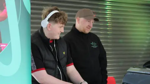Kay Davidson/BBC Harry, a teenage boy with short blonde hair stands on the right side of an adult man. Harry is wearing white over‑ear headphones, a black T‑shirt with red cuffs and a black body warmer. Harry is wearing a brown cap and a black zip-up fleece. They are both slightly smiling, their heads are turned slightly to the right looking at a silver open laptop. They are standing behind the desk, positioned in front of a closed shopfront with its shutters down. To the left there is a tall blue pop‑up banner.