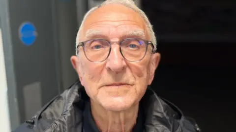 Geoff Hallewell with short white hair wearing grey glasses and a black jacket and polo top. He looks impassively.