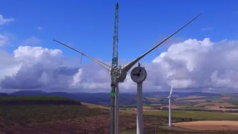 Huntly Development Trust Close up of a wind turbine wings being placed onto its mast. There are fields behind the large tall white stand. 