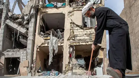 A man in a white turban and black clothes brushes the area in front of a bombed out building