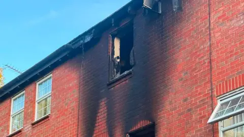 Paul Graham The outside of a home that has been severely burned by a fire. The home is made from red brick, a window is no longer there, and smoke damage has risen up the outside wall. A white window is open to the right and two shut windows to the right. 