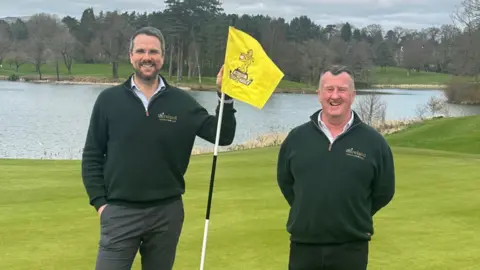 18Ireland Mark Faloon, a man with dark grey hair and beard, wearing a green top, beside a yellow golf hole flag. Beside him to the right, is Gary O'Hara, a man with dark grey hair wearing a green top. They are on the green of a golf course and there is a lake in the background.