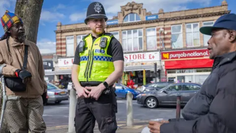 wmp A policeman stood in between two men, having a conversation with them. The man on the left is stood up but leaning on a crutch. He wears a blue, yellow and red patch work hat, brown zip jacket and black and brown patterned trousers. The man stood to the right of the officer is seated, wearing a blue hat, black puffer jacket and holding a plastic cup.