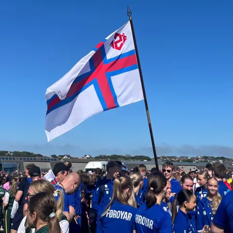 The Faroe Islands flag blowing in the wind, surrounded by athletes