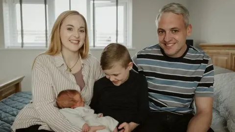 Hayley and Mike with their children George and Hope. The family are sitting on a bed and are smiling at the camera. 