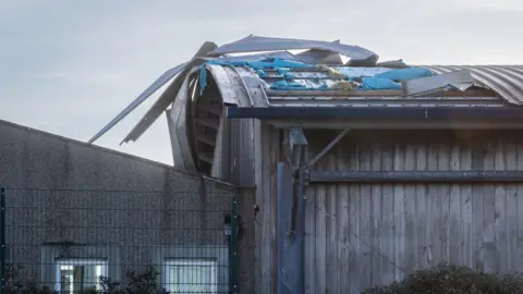 The image shows a building with severe roof damage. The structure appears to be made of wood and metal, with a curved metal roof that has been torn and twisted. Large sheets of metal have peeled back and are bent upwards, exposing insulation material underneath. The damage looks consistent with strong winds, possibly from a storm. The walls of the building are intact, and there is a metal fence in the foreground. Two small windows are visible on the left side of the image, and some bushes are growing near the base of the building.